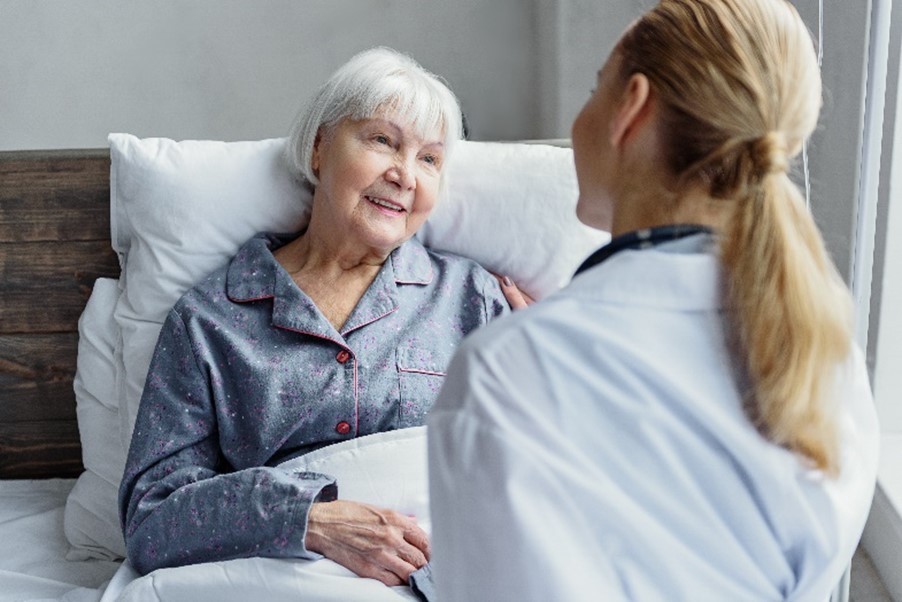 Seniorin im blauen Schlafanzug liegt in einem weißen Bett und lächelt eine Pflegerin vor sich an, die mit dem Rücken zur Kamera steht.