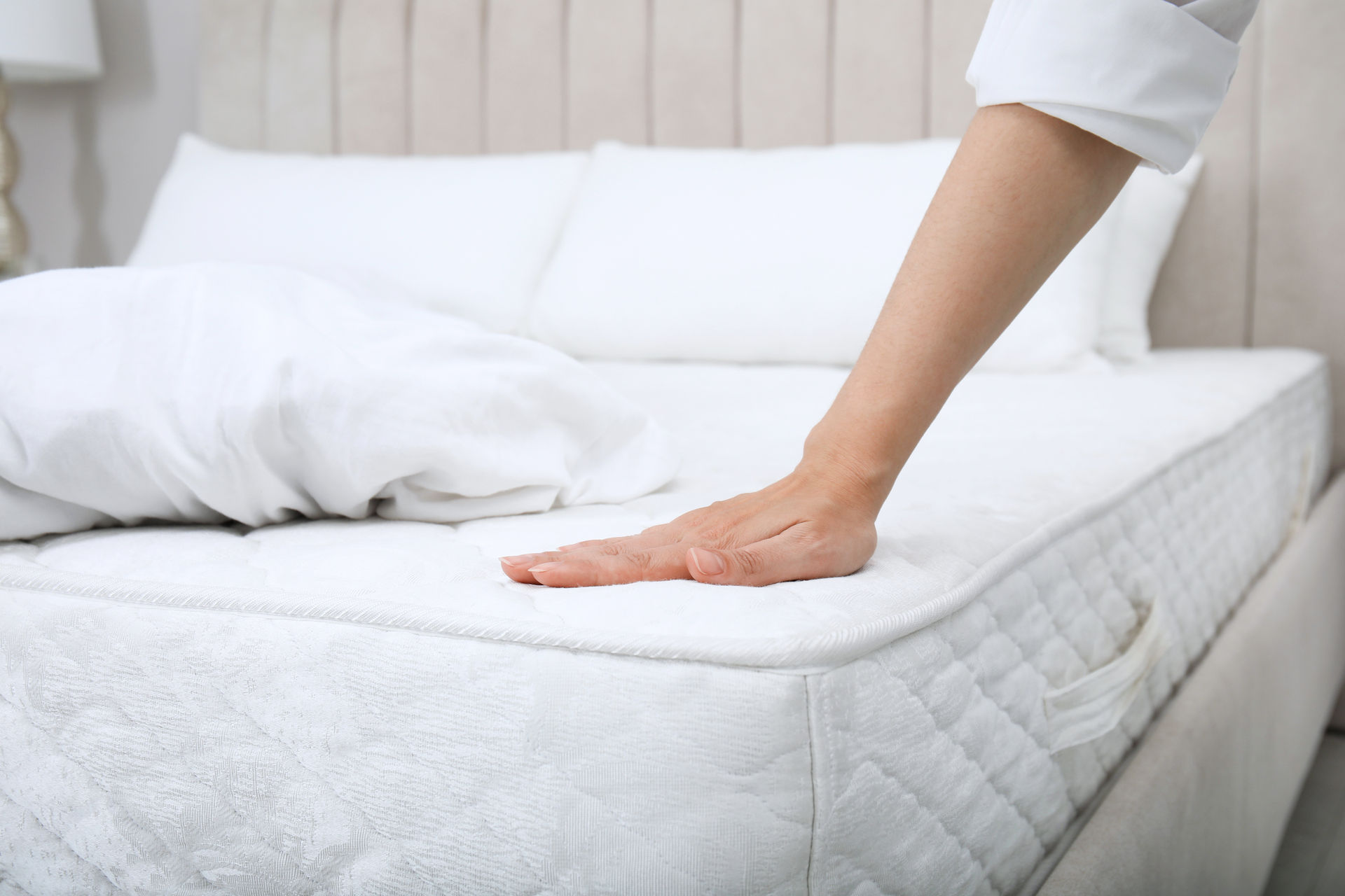 matratzenreinigung-hotel-hotelbett-mit-weißer-matratze-und-weißer-bettwaesche-wo-eine-hand-drueber-faehrt Ein Bett ist mit einer weißen Matratze. Kissen und einer Decke ausgestattet. Auf der Matratze ruht eine Hand mit sichtbarem Unterarm.