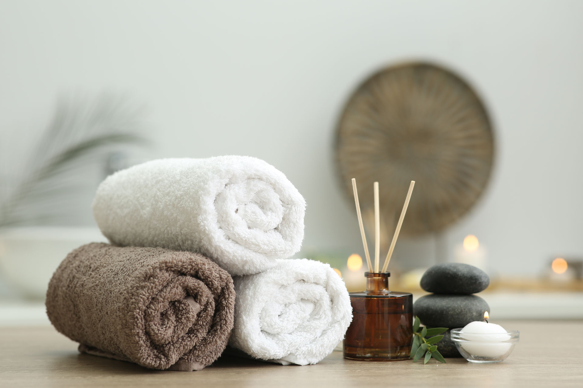 wellness-textilien-gerollte-handtuecher-mit-weitere-wellness-utensilien Gerollte Handtücher, Aroma-Diffuser, Massagesteine und brennende Kerze liegen und stehen auf einem Holztisch.