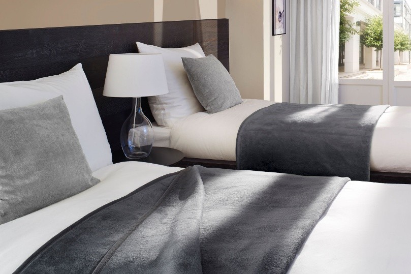 Betten im Hotelzimmer sind vorbereitet. Auf den Bettdecken liegen graue Kuscheldecken.