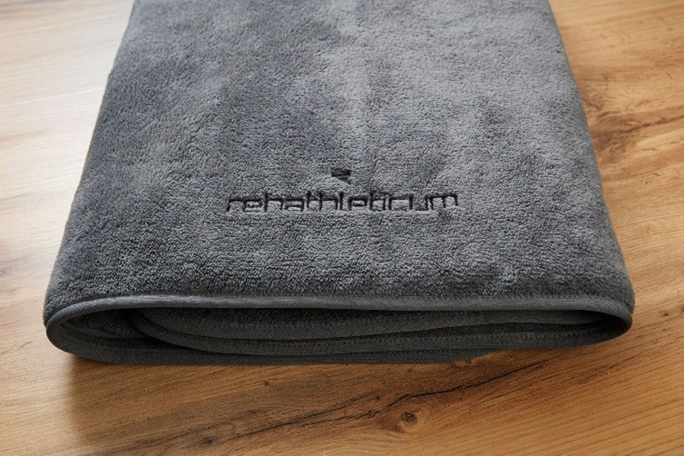 Graues Frottee-Handtuch mit schwarzer Logo-Stickerei „rehathleticum“ auf Holzoberfläche.