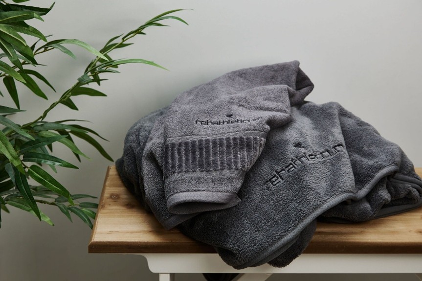 Zusammengelegte graue Frottee-Handtücher mit Logo-Stickerei „rehathleticum“ auf Holzoberfläche.