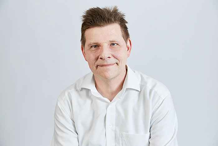 Lächelnder Mann mit kurzen braunen Haaren, trägt ein weißes Hemd, vor neutralem Hintergrund.