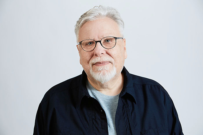 Lächelnder älterer Mann mit grauen Haaren, Bart und Brille, vor neutralem Hintergrund.