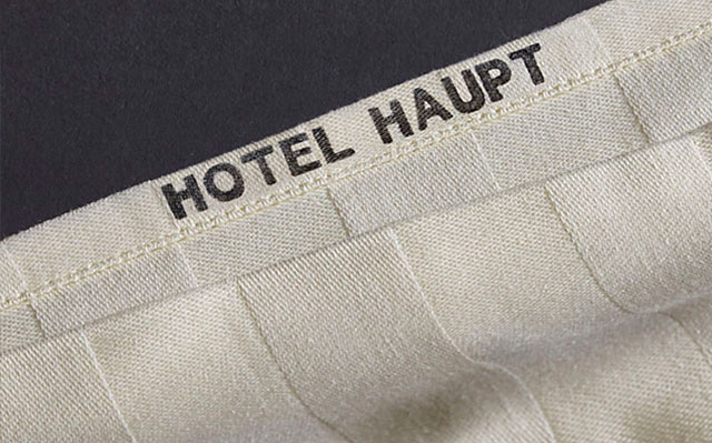 Ein beigefarbener Stoff mit einer schwarzen Einstempelung mit dem Text "Hotel Haupt" in einem dunklen Hintergrund.