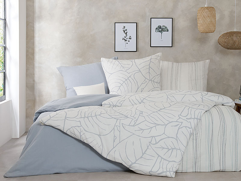 Crêpe-Bettwäsche Bio-Baumwolle farbig In einem Zimmer steht ein großes Doppelbett. Auf dem Bett liegen Bettdecken und Kissen, die mit grauer und gemusterter Bettwäsche bezogen wurden.