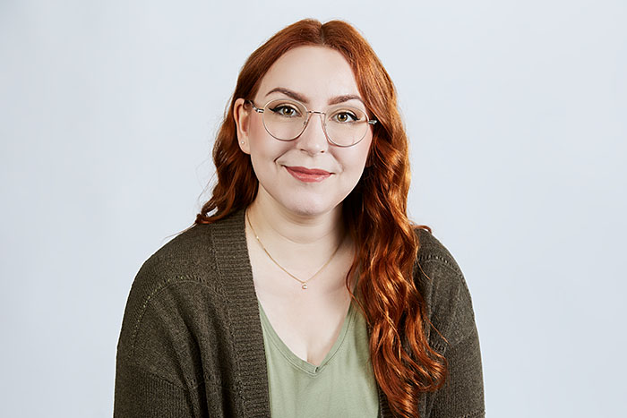 Lächelnde Frau mit langen roten Haaren und Brille, grünem Oberteil mit Strickjacke, vor neutralem Hintergrund.