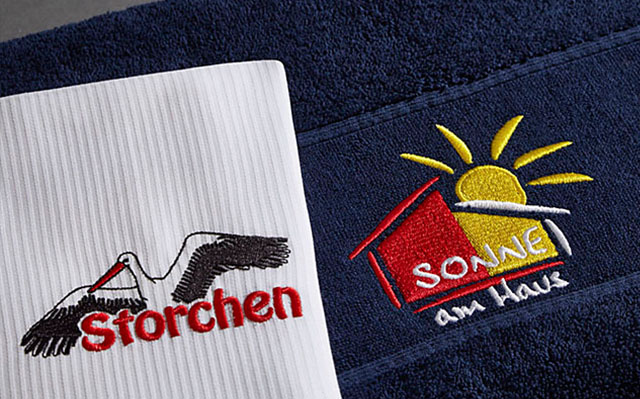 Handtücher zeigen gestickte Logos, eines mit einem Storch, das andere mit Sonne und Haus.
