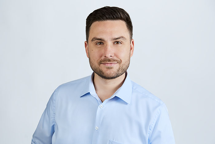 Lächelnder Mann mit kurzem dunklem Haar und Bart, hellblauem Hemd, vor neutralem Hintergrund.