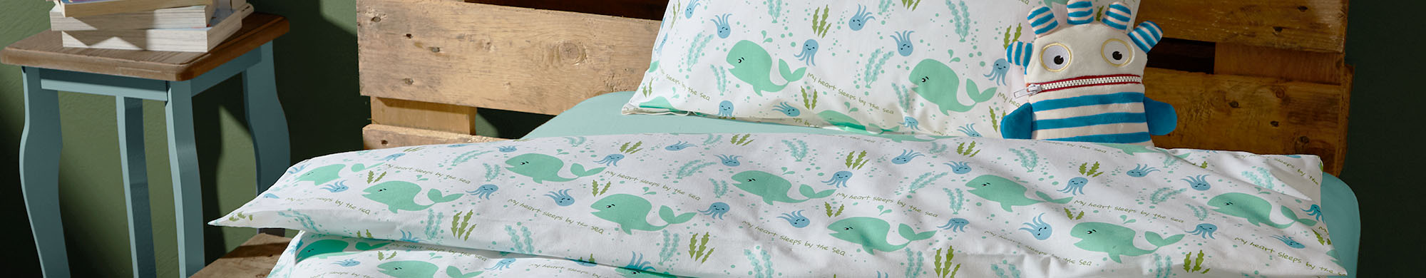Bettzeug mit Wal-Motiven befindet sich auf einem Holzrahmen; Plüschtier sitzt daneben.