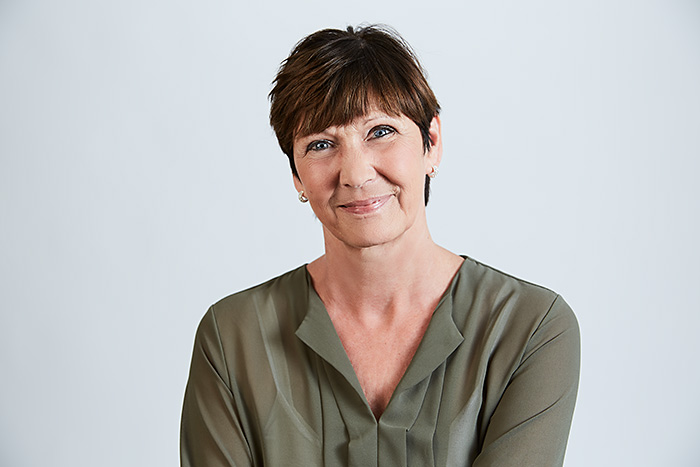 Lächelnde Frau mit kurzen braunen Haaren, trägt eine olivgrüne Bluse, vor neutralem Hintergrund.