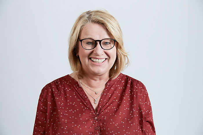 Lächelnde Frau mit blonden Haaren, Brille und roter Bluse mit Muster, vor neutralem Hintergrund.