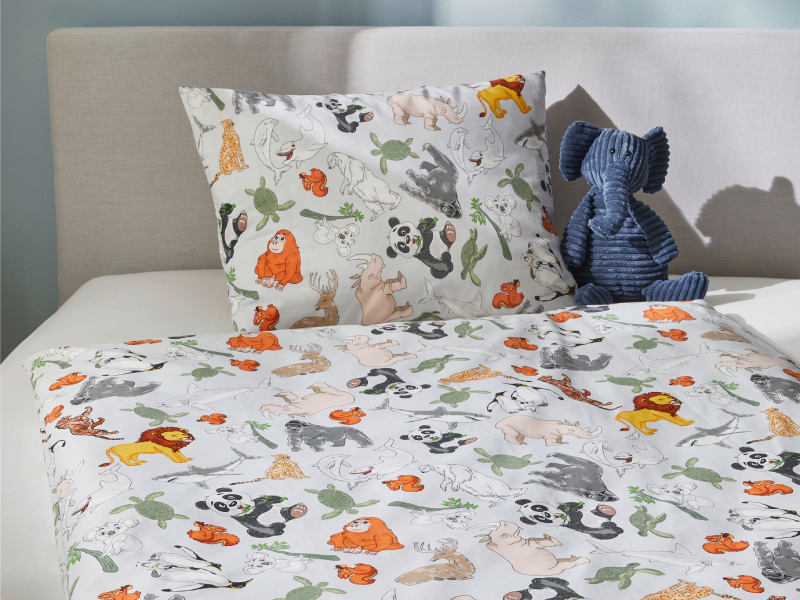 Bett mit farbenfroher Kinderbettwäsche im Tierpark-Design, inklusive Tieren wie Elefant und Giraffe in einem hellen Kinderzimmer.